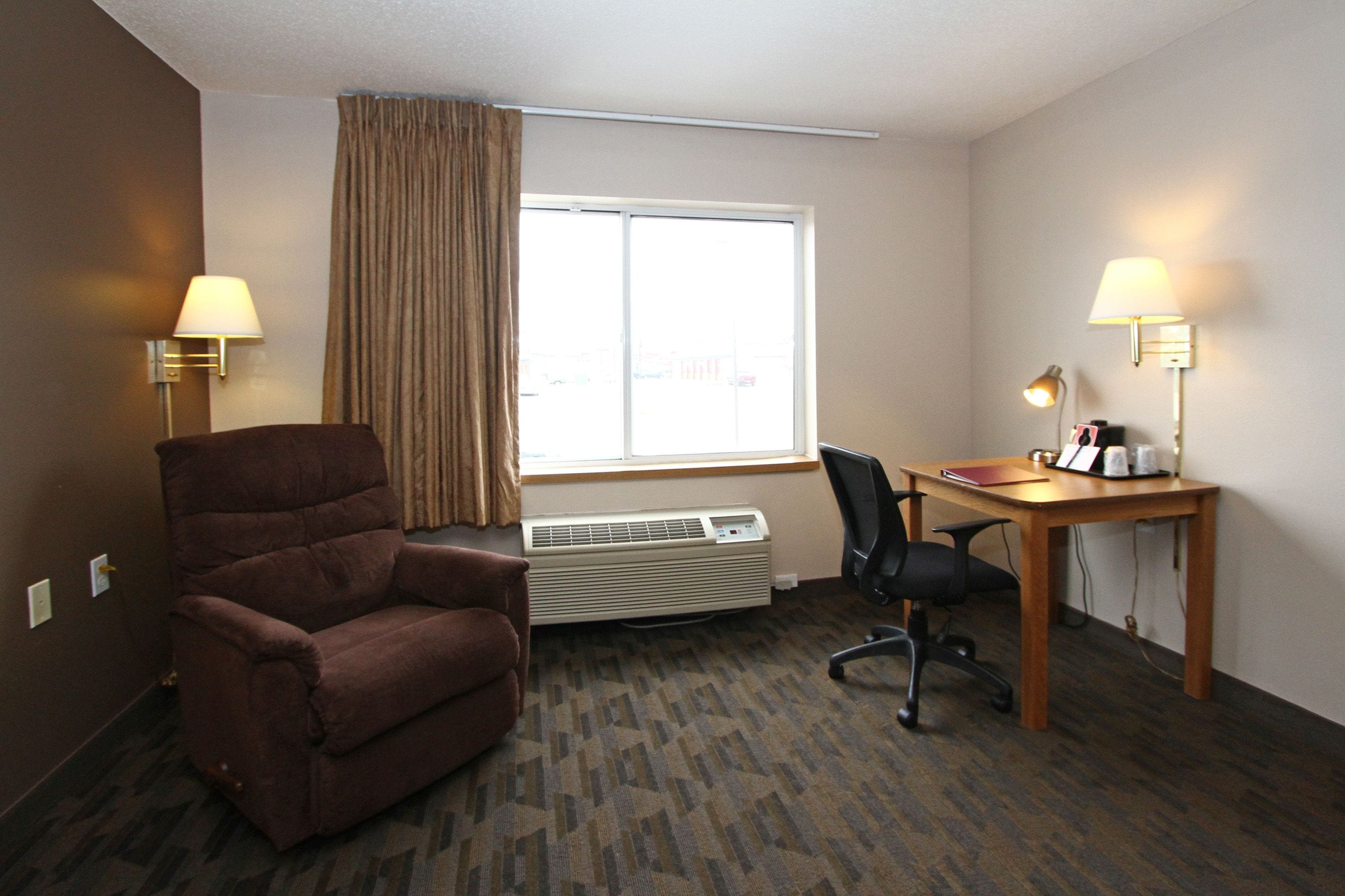 A hotel room with a brown armchair, a desk with a lamp, a chair, and a large window with curtains.