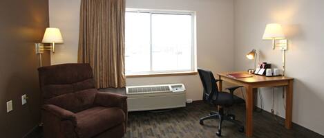A hotel room with a brown armchair, a desk with a lamp, a chair, and a large window with curtains.