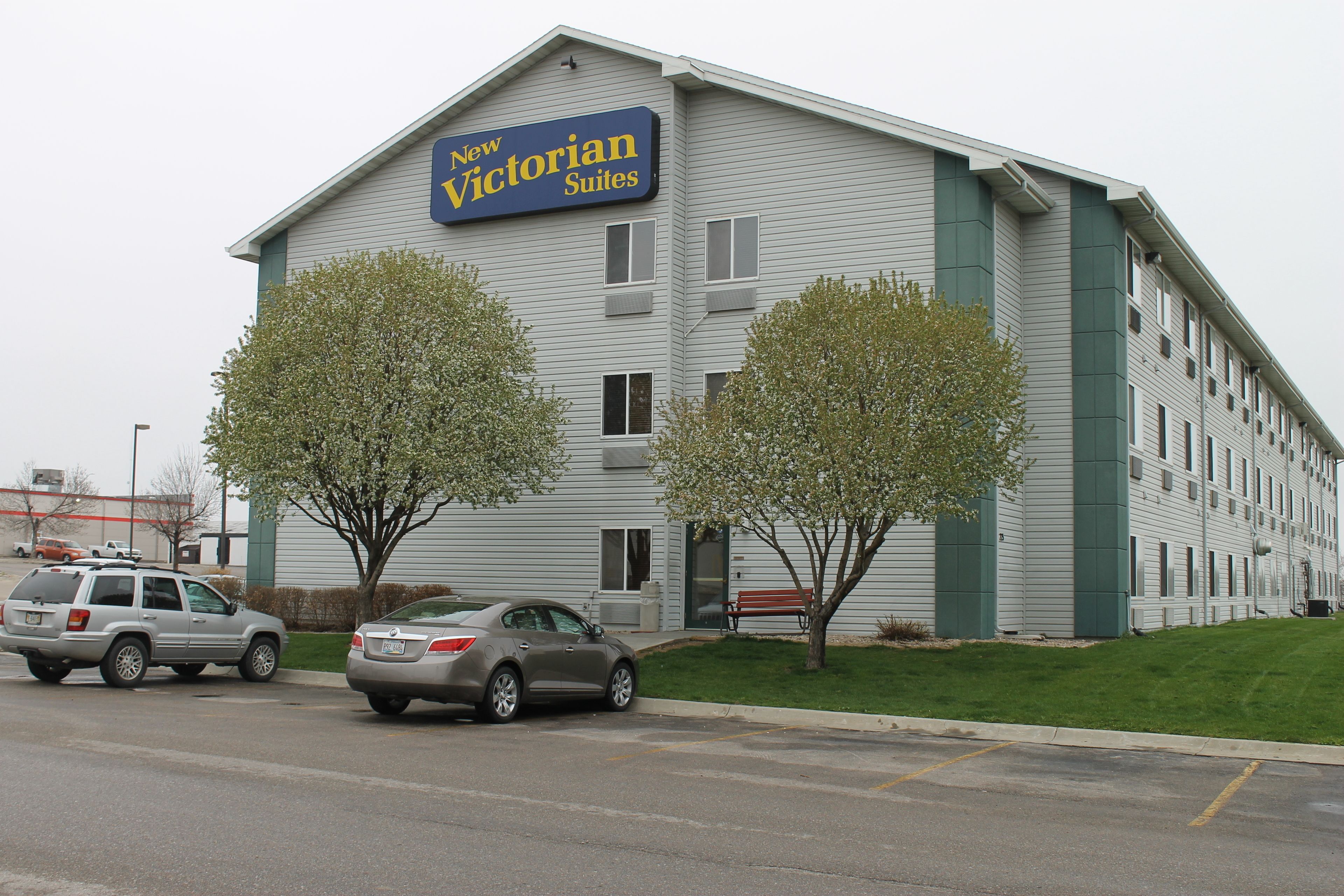 A building with a sign reading "New Victorian Suites." There are two cars parked in front, and two trees are visible in front of the building.