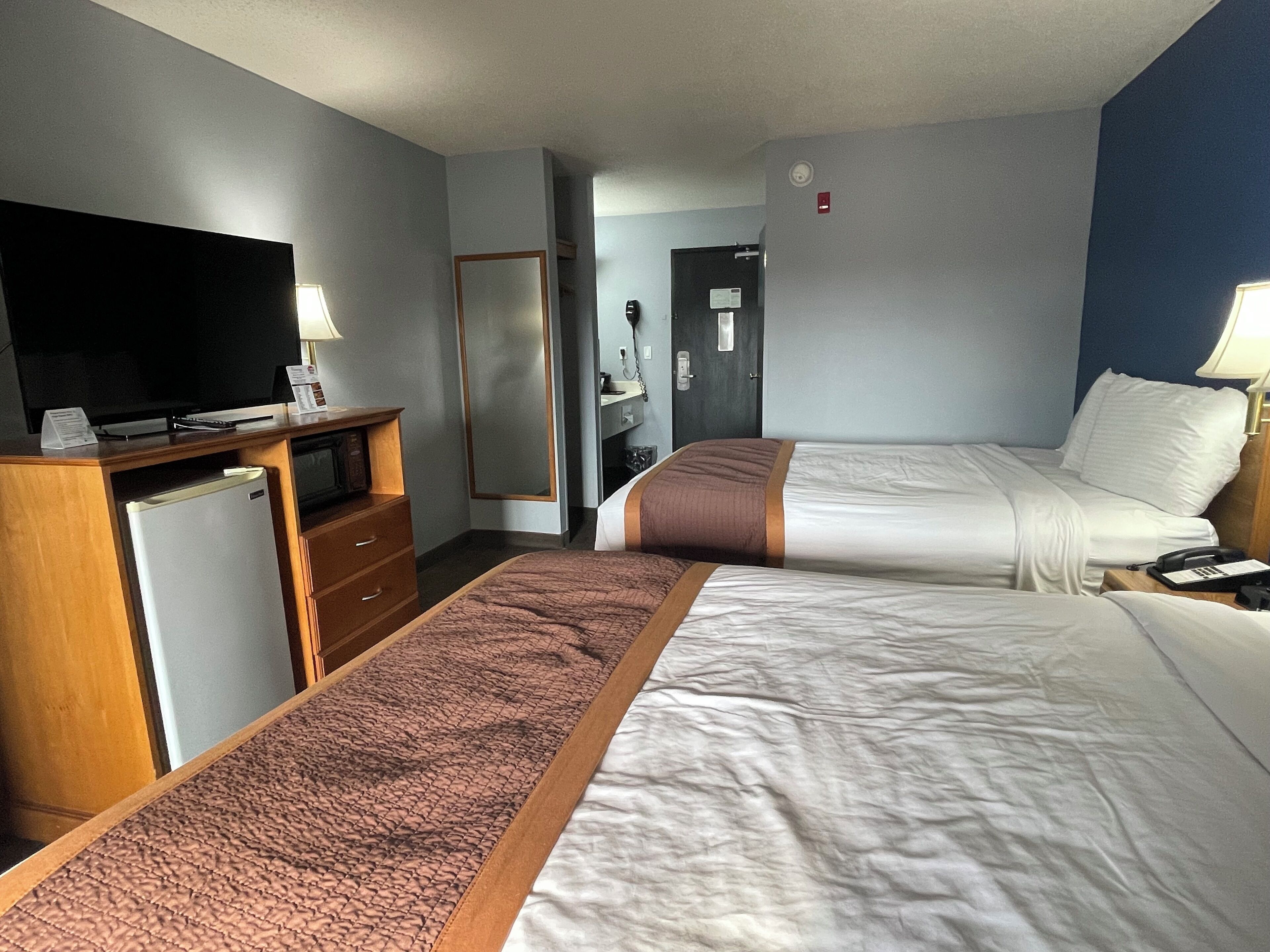 A hotel room with two beds, a desk with a television, a refrigerator, and a mirror.
