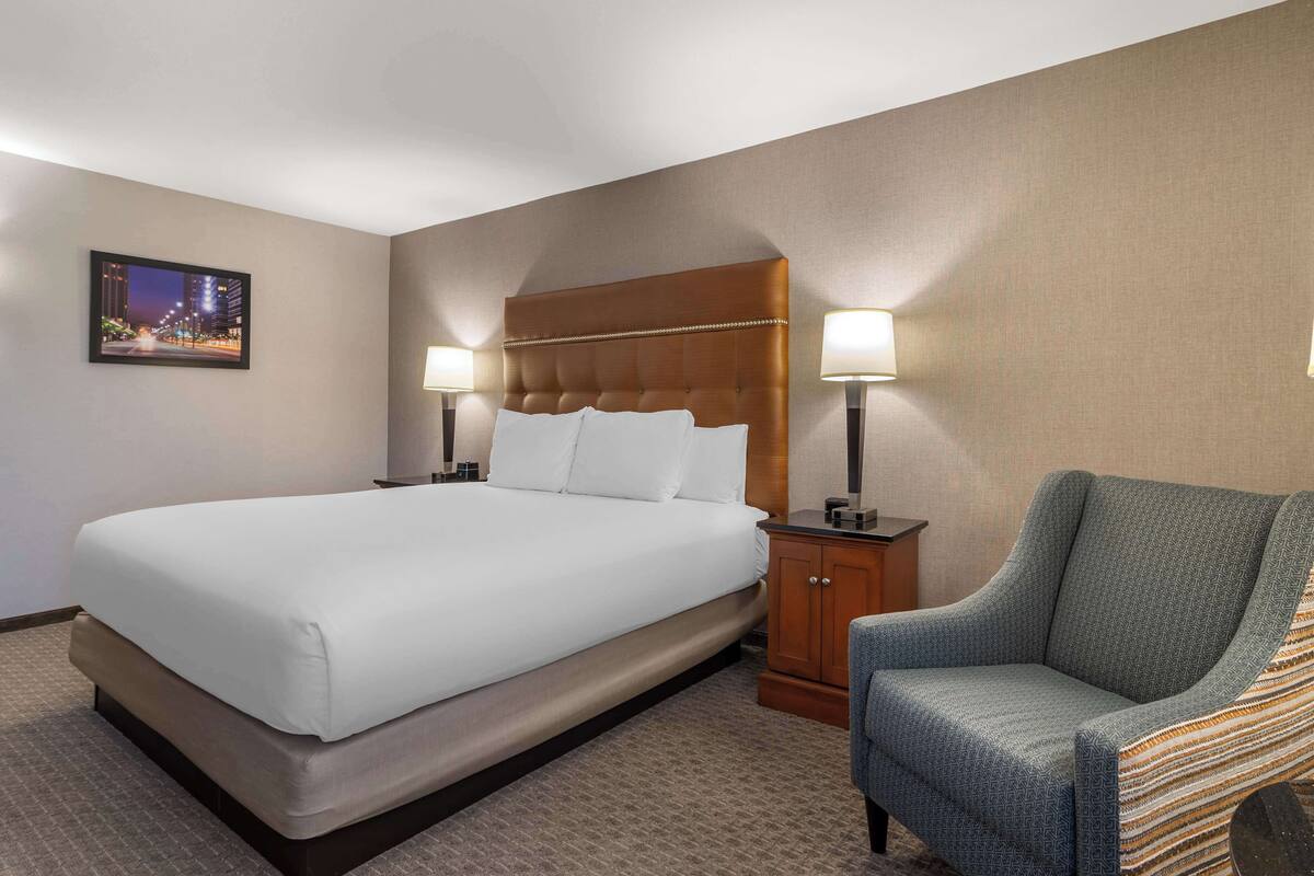 A hotel room with a large bed, two bedside lamps, a chair, and a framed picture on the wall.