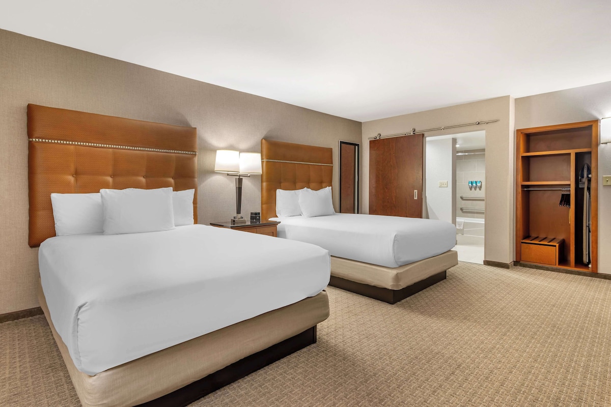 A hotel room with two beds, a nightstand, a lamp, a closet, and a bathroom visible through an open door.