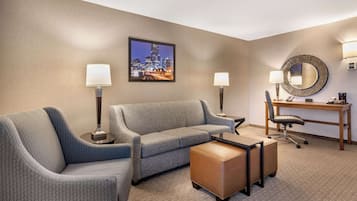 A hotel room with a grey sofa, a small ottoman, a desk with a chair, a round mirror, and a framed cityscape picture on the wall.
