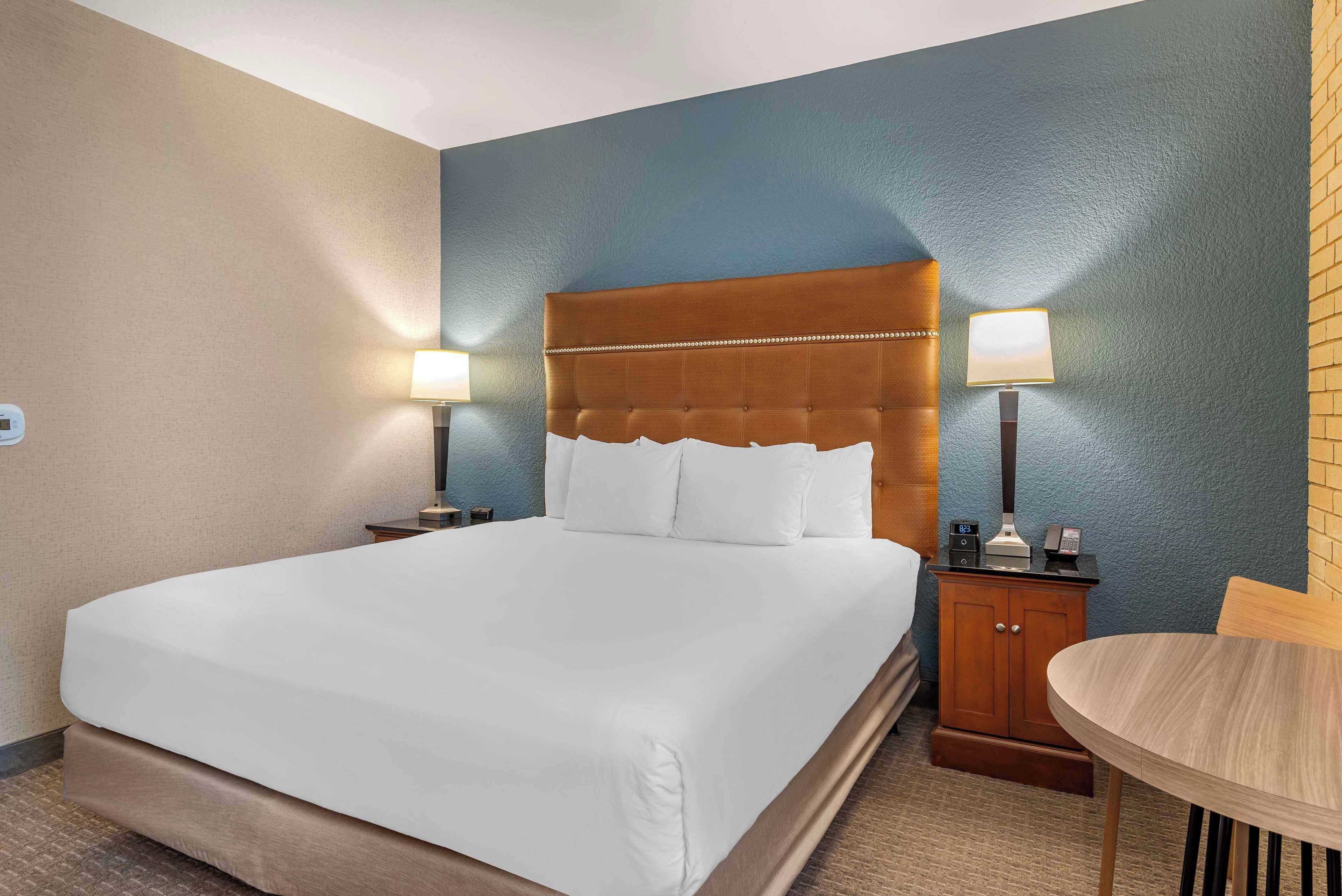 A hotel room with a large bed, two bedside tables with lamps, a wooden nightstand, and a small table with a chair.
