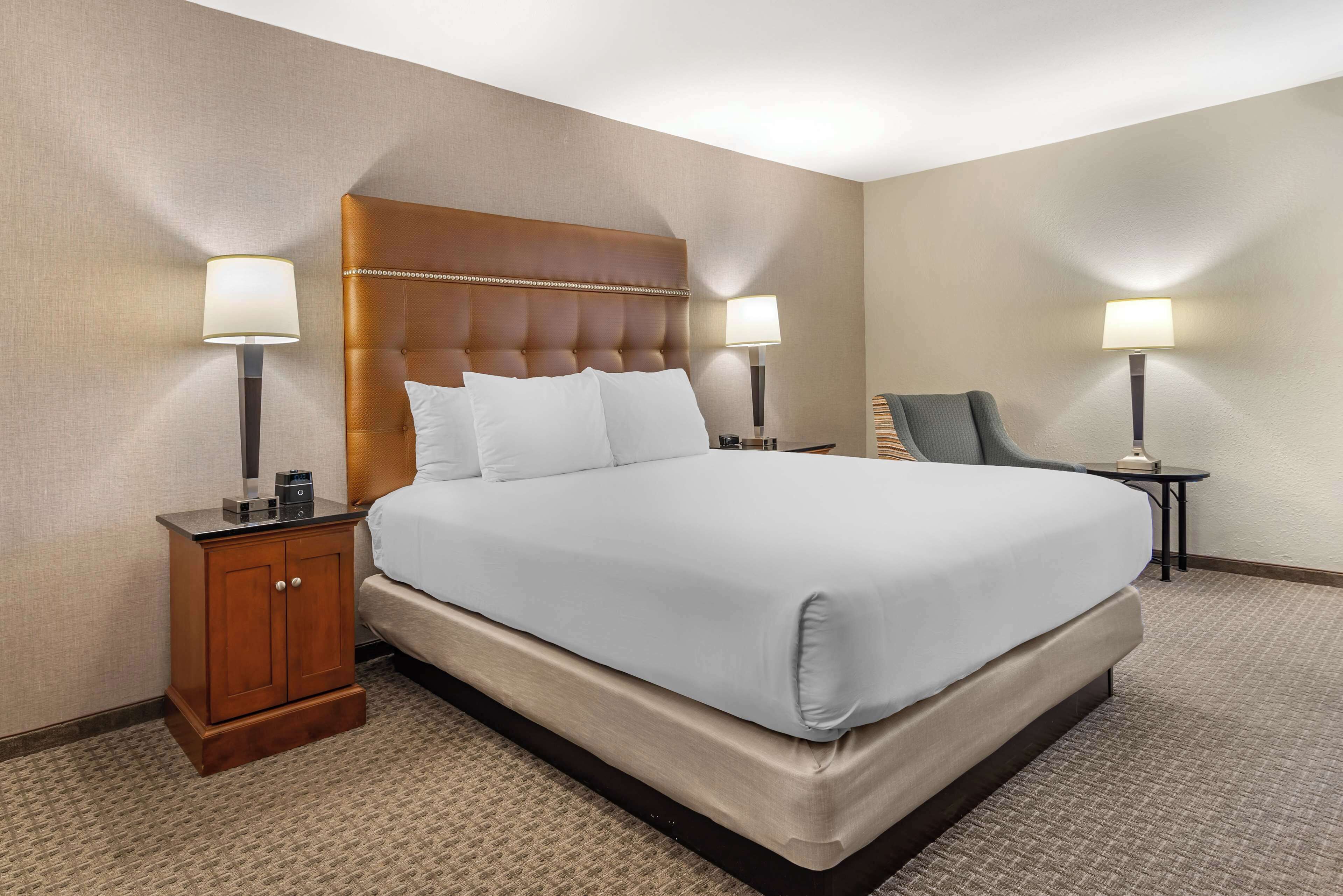 A hotel room with a large bed, two bedside tables with lamps, a wooden nightstand, and a carpeted floor.