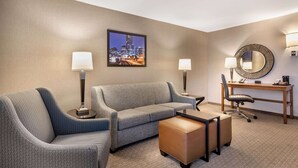 A hotel room with a grey sofa, a small ottoman, a desk with a chair, a round mirror, and a framed cityscape picture on the wall. - Drury Inn & Suites Atlanta Airport (Atlanta)