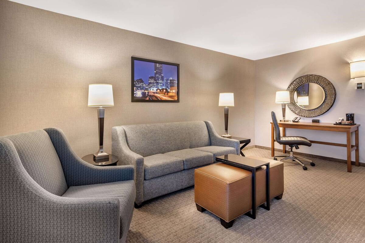 A hotel room with a grey sofa, a small ottoman, a desk with a chair, a round mirror, and a framed cityscape picture on the wall.