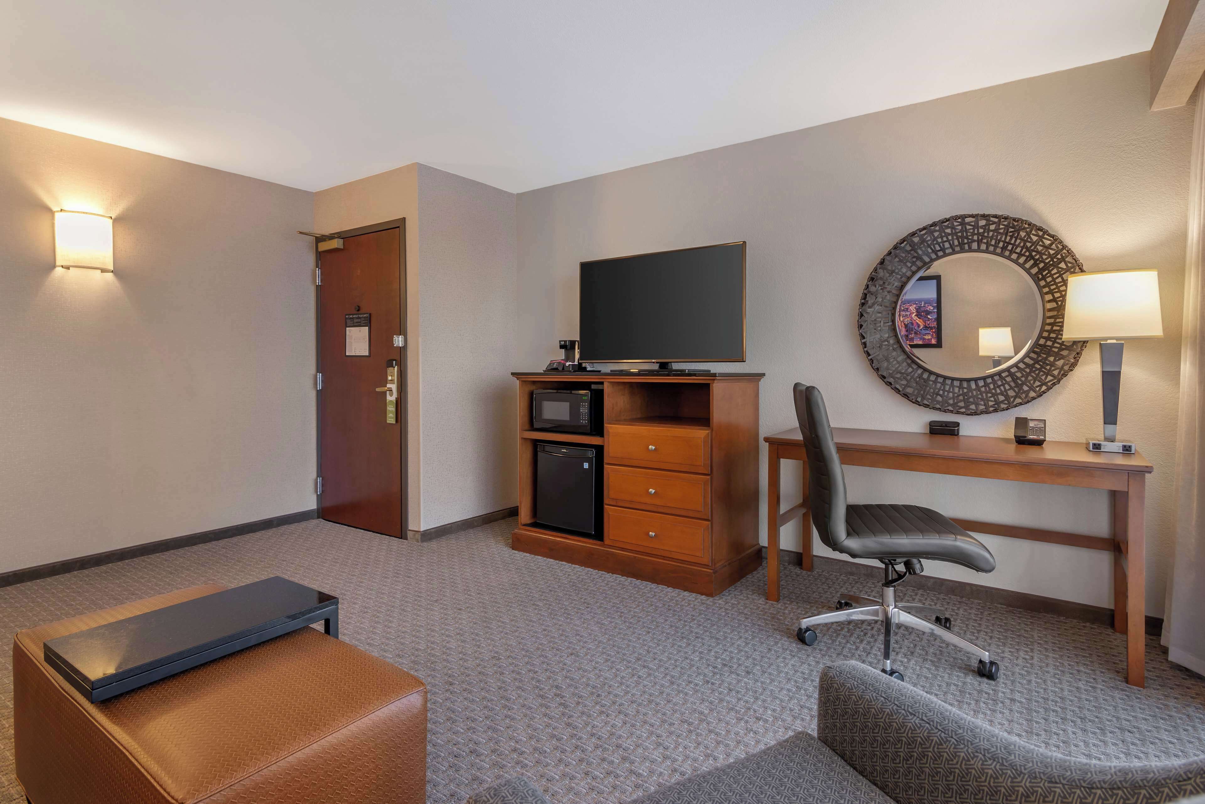 Habitación de hotel con escritorio, silla, televisión, minibar y una pequeña zona de estar.