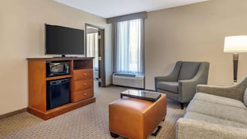 Habitación de hotel con televisión de pantalla plana, un armario de madera con microondas, un sofá, una mesa de centro y un amplio ventanal con cortinas.