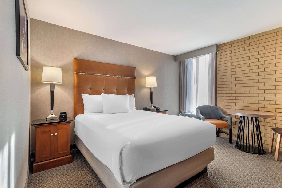 A hotel room with a large bed, two bedside tables with lamps, a small table, a chair, and a brick wall.