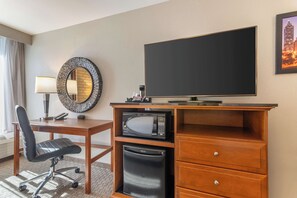 A hotel room with a wooden desk, a black office chair, a microwave, a small refrigerator, a flat-screen TV, a mirror, and a window with curtains.