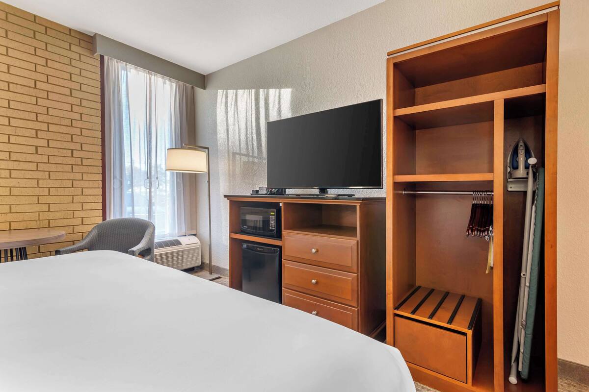 A hotel room with a bed, a desk with a lamp, a television, a microwave, a small refrigerator, a closet, and a ironing board.