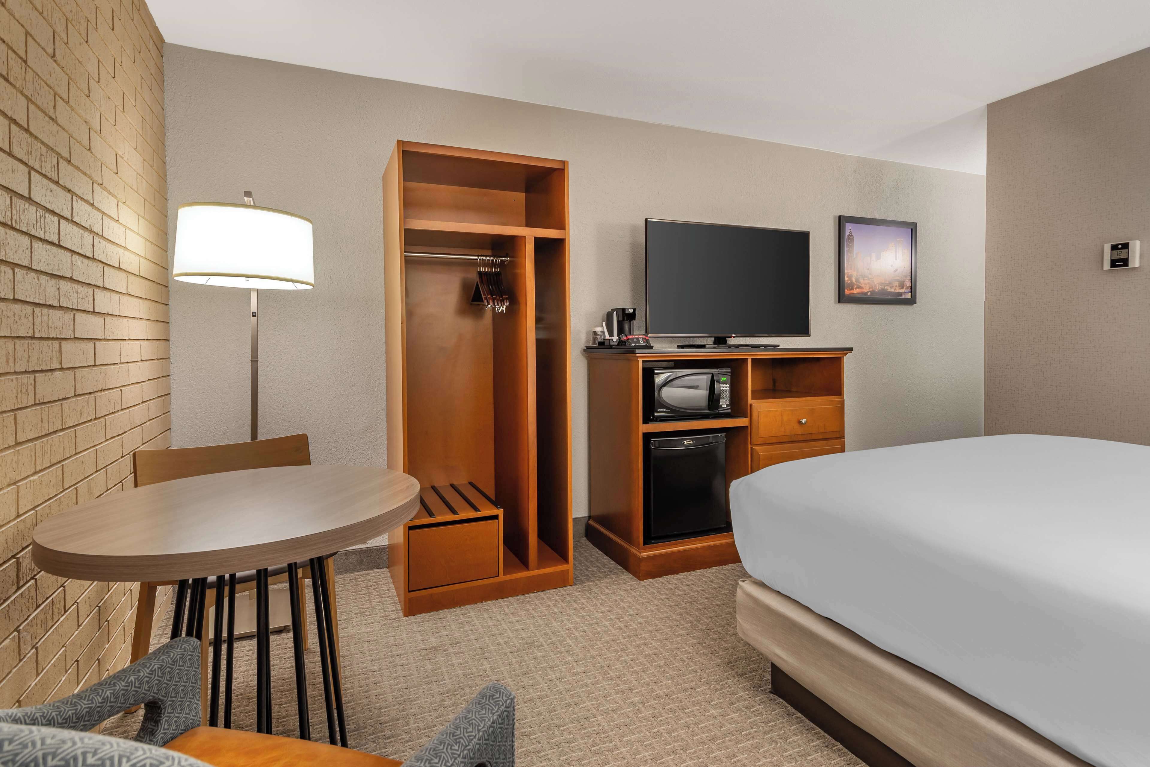 A hotel room with a bed, a desk with a lamp, a television, a wardrobe, and a small kitchenette.