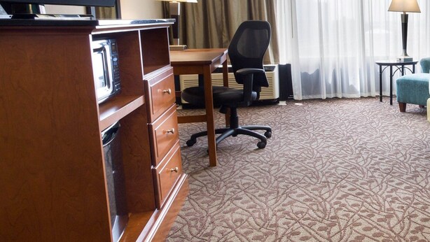 A hotel room with a flat-screen TV, a wooden dresser, a desk with a chair, a lamp, and a large window with curtains. - Drury Inn & Suites Atlanta Airport (Atlanta)