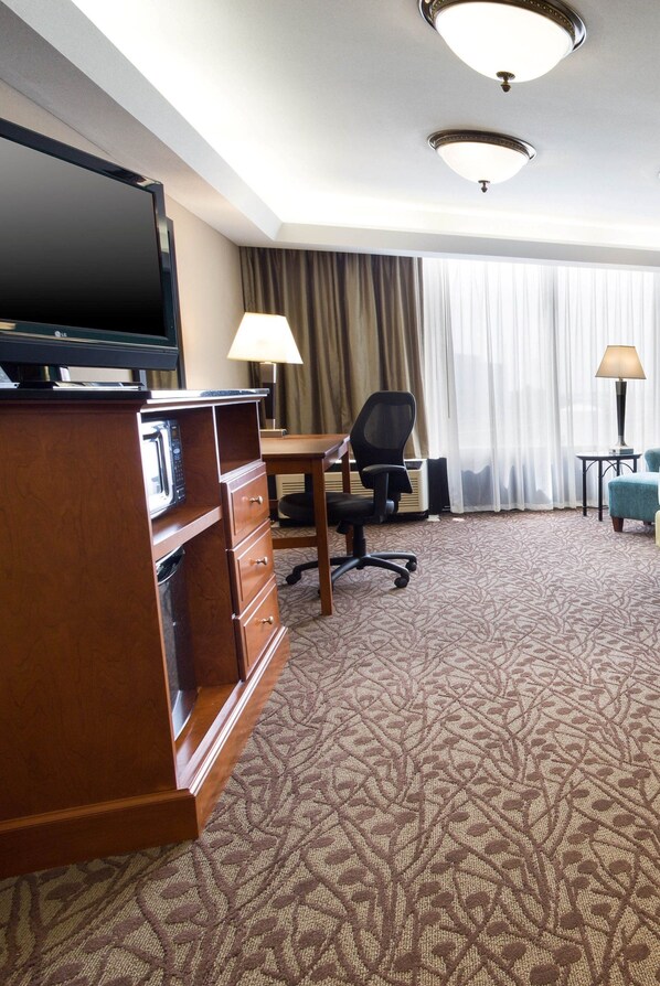 A hotel room with a flat-screen TV, a wooden dresser, a desk with a chair, a lamp, and a large window with curtains. - Drury Inn & Suites Atlanta Airport (Atlanta)