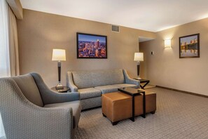 A hotel room with a grey sofa, a brown ottoman, two floor lamps, and two framed pictures on the walls. - Drury Inn & Suites Atlanta Airport (Atlanta)