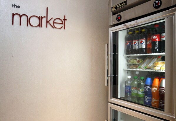 Un refrigerador lleno de bebidas y aperitivos, un letrero en la pared que dice 'the market', y una pantalla digital sobre el refrigerador.
