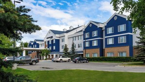 A multi-story hotel with a blue and white exterior, a parking lot with several cars, and surrounding greenery. - SpringHill Suites Anchorage Midtown (Anchorage)
