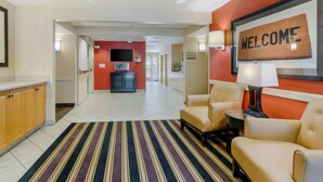 A hotel lobby with a striped carpet, a reception desk, a 'WELCOME' sign, and a TV on the wall. - Extended Stay America Suites Pleasanton Chabot Dr (Pleasanton)