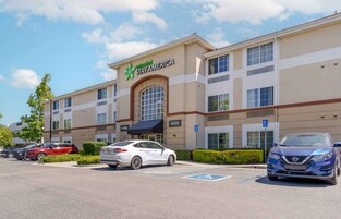 Un hotel StayAmerica con fachada moderna, entrada al vestíbulo y un estacionamiento con varios coches.