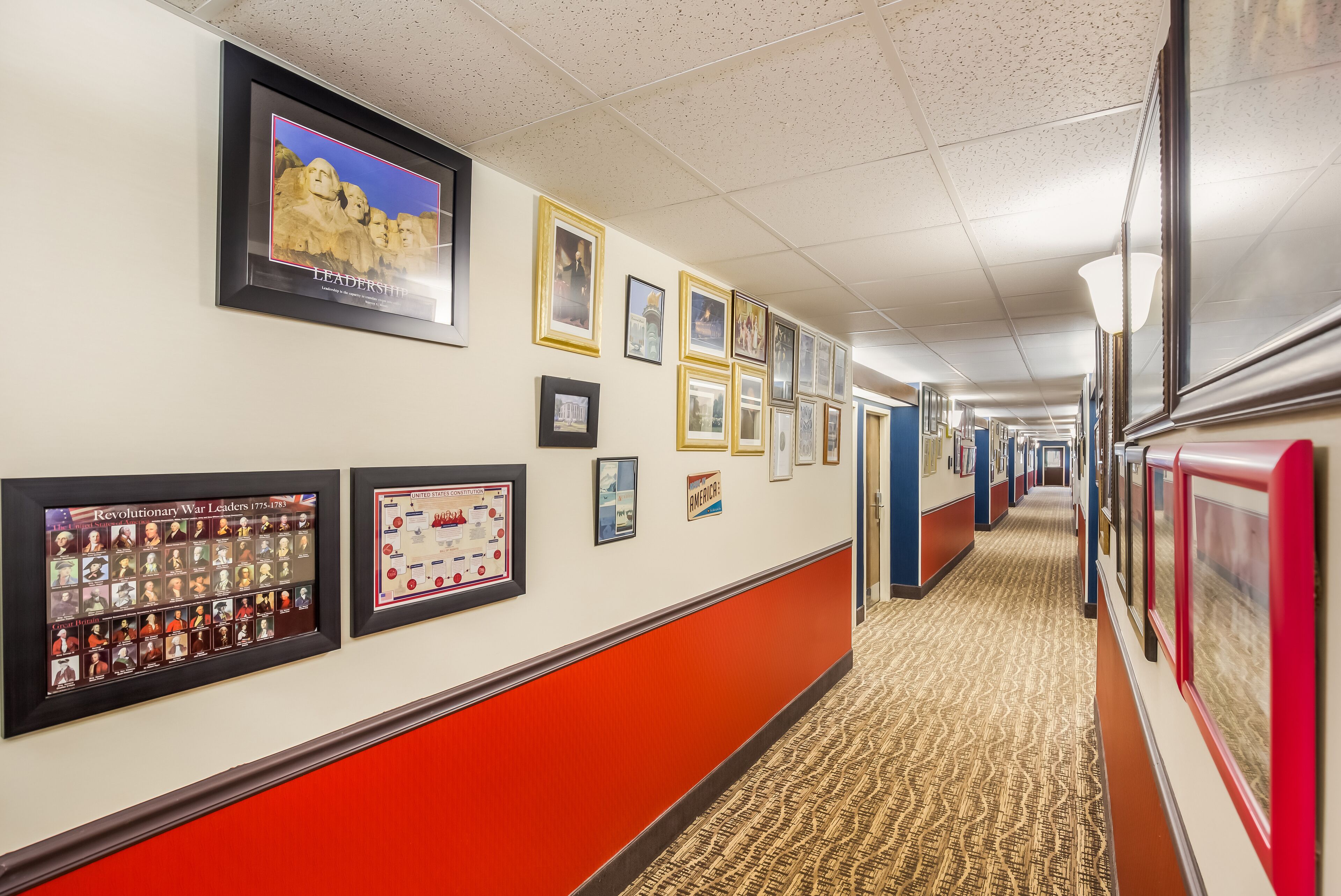 A hallway with framed pictures on the walls, a carpeted floor, and a red and white wall design.