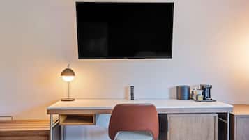 Una habitación de hotel moderna con una televisión de pantalla plana montada en la pared, un escritorio con lámpara, una silla y una cafetera.