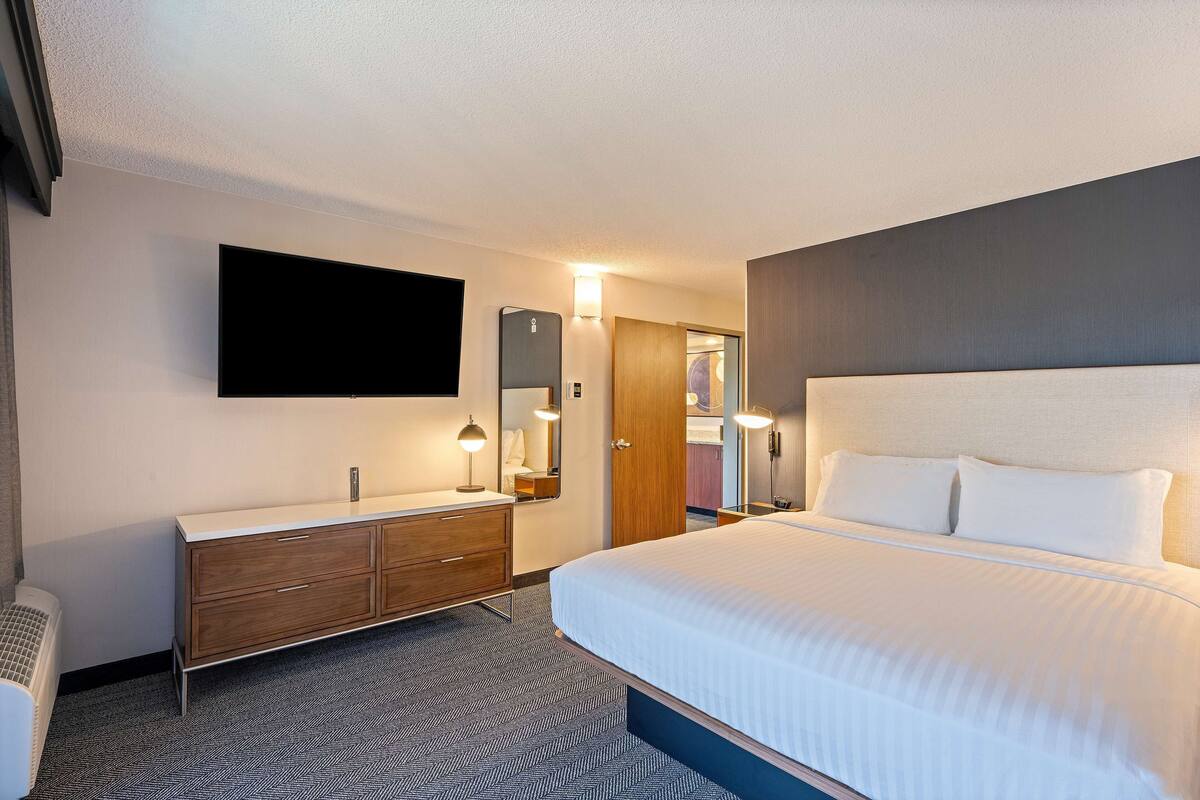 A hotel room with a large bed, a flat-screen TV mounted on the wall, a wooden dresser with drawers, a mirror, and a door leading to a bathroom.