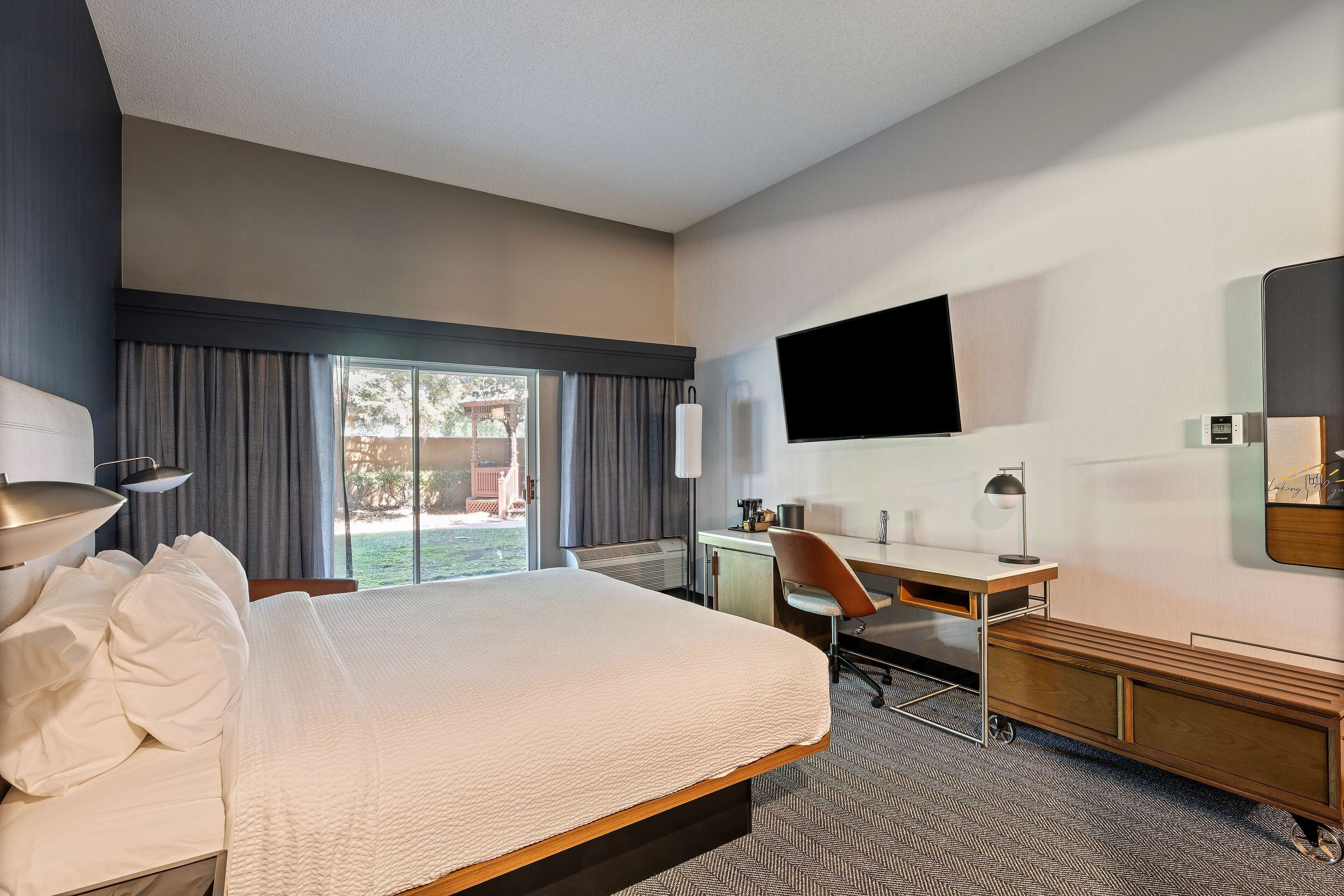 Habitación de hotel con una cama grande, un escritorio, una silla, un televisor montado en la pared y una ventana con cortinas.