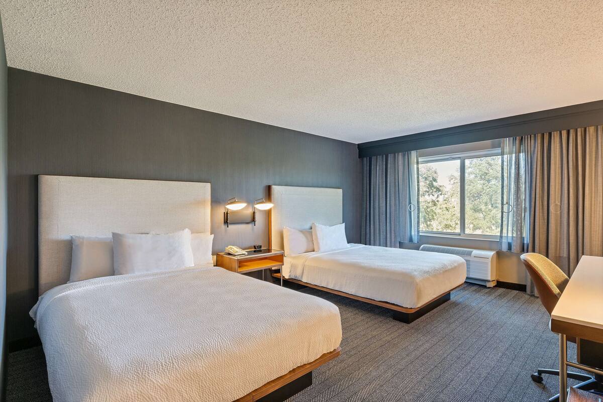 A hotel room with two beds, a desk, and a chair. There is a window with curtains and an air conditioning unit.