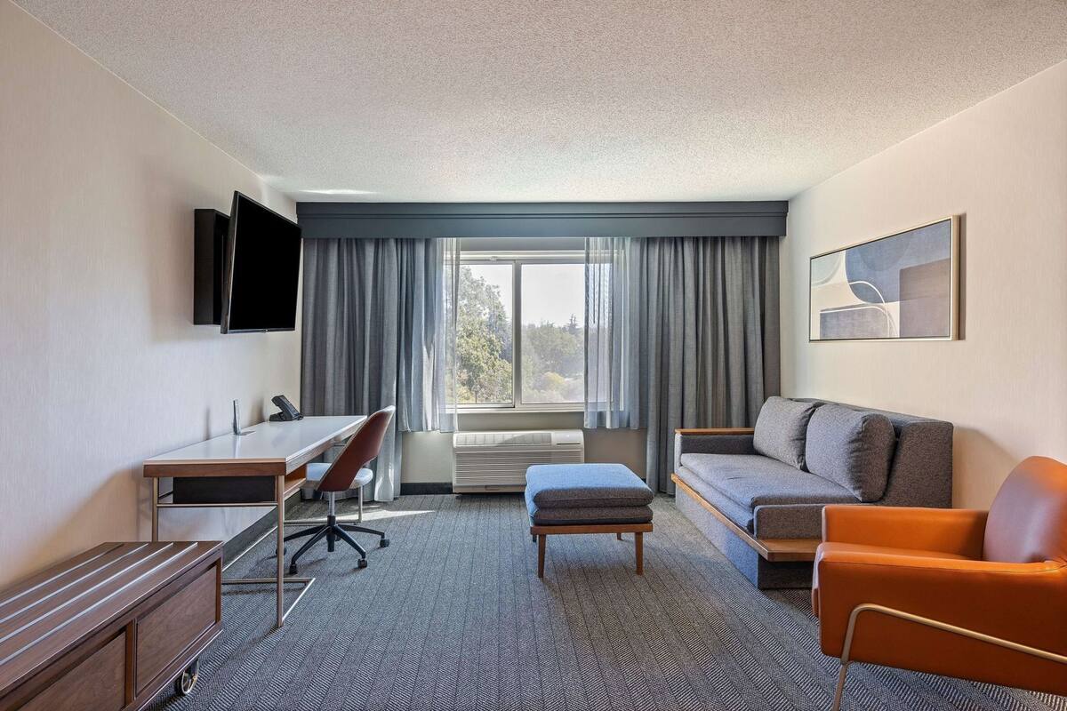 A hotel room with a desk, chair, sofa, ottoman, and a TV mounted on the wall.