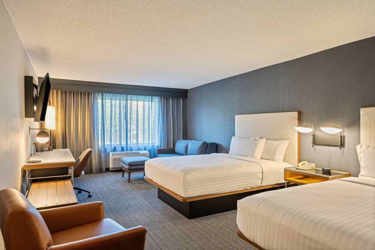 A hotel room with a large bed, a desk with a lamp, a chair, and a bedside table with a phone.