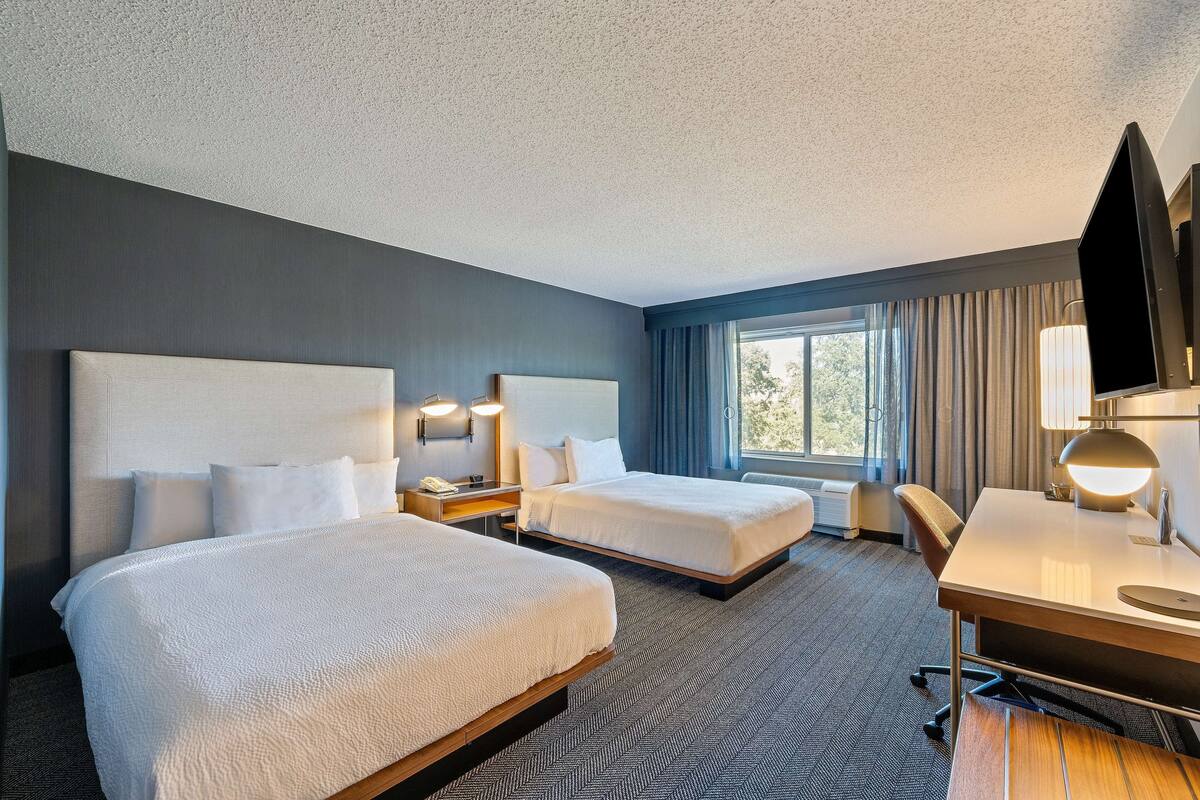 A hotel room with two beds, a desk, a chair, a TV, and a window with curtains.