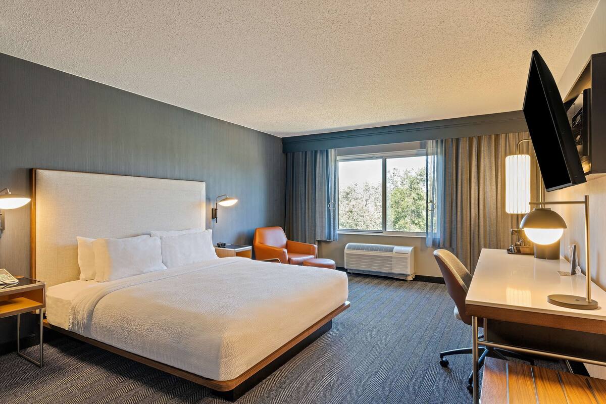 A hotel room with a large bed, a desk, a chair, a TV, and a window with curtains.