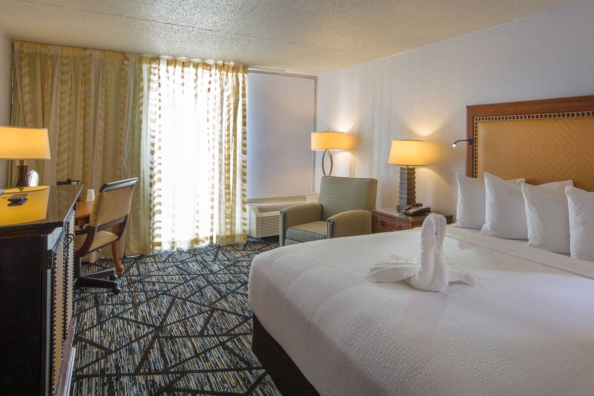 A hotel room with a large bed, two bedside lamps, a desk with a chair, and a patterned carpet.