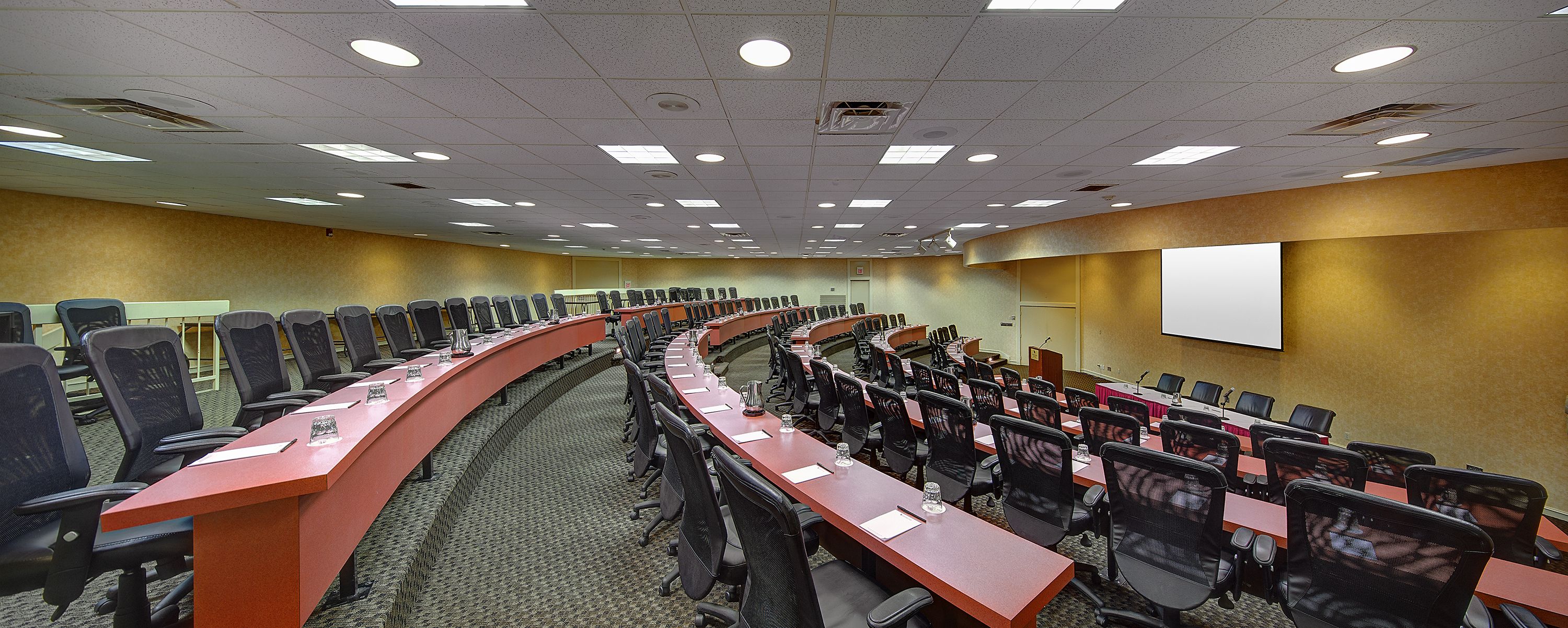 A conference room with a long table and rows of chairs, equipped with microphones and a projector screen.