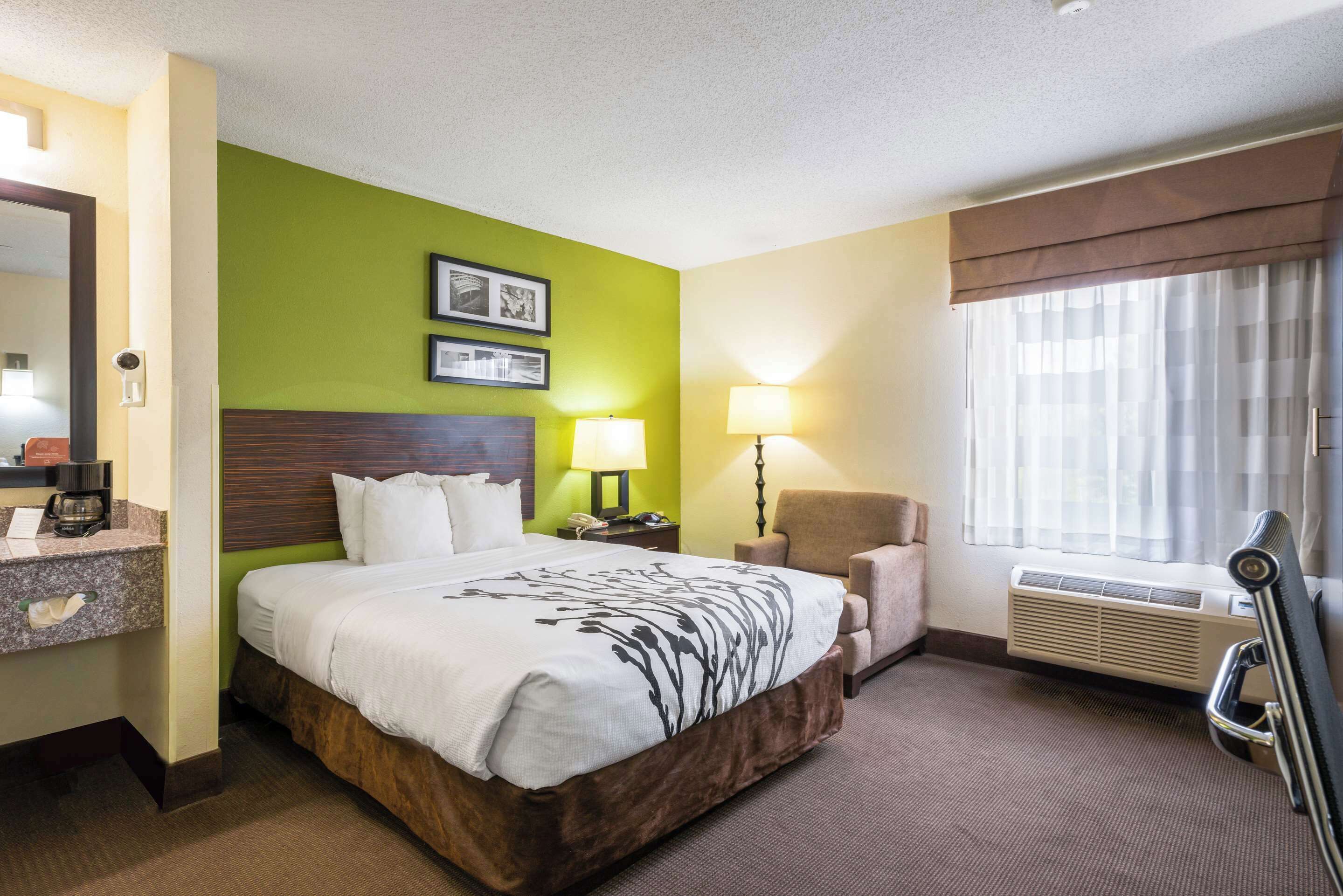 A hotel room with a large bed, a desk with a lamp, a chair, a window with curtains, and a wall-mounted air conditioner.