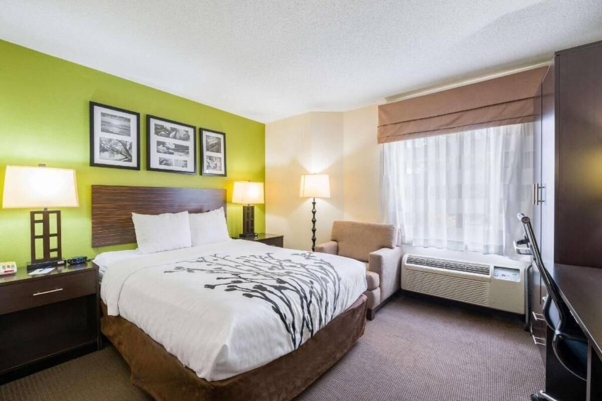 Ein Hotelzimmer mit einem Bett, Nachttischlampen, einem Sessel, einem Fenster mit Vorhängen und einer Wand-Klimaanlage.