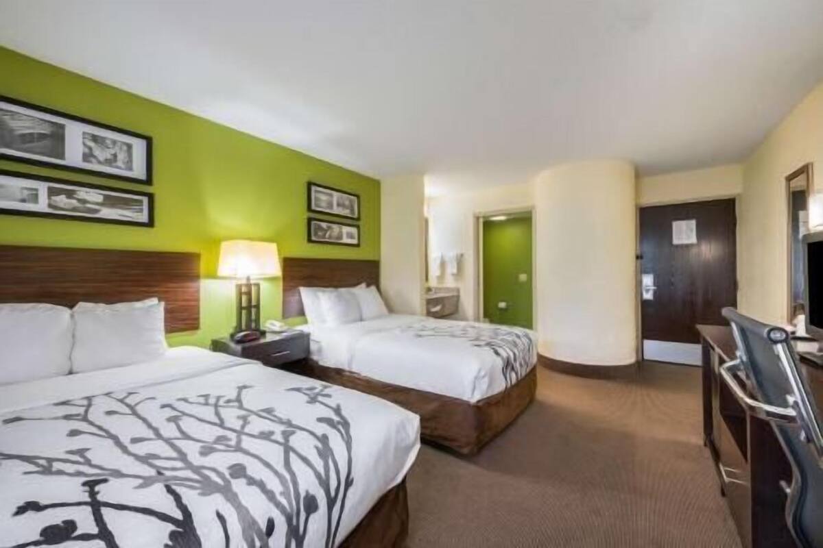A hotel room with two beds, a desk, a chair, a lamp, and framed pictures on the wall.