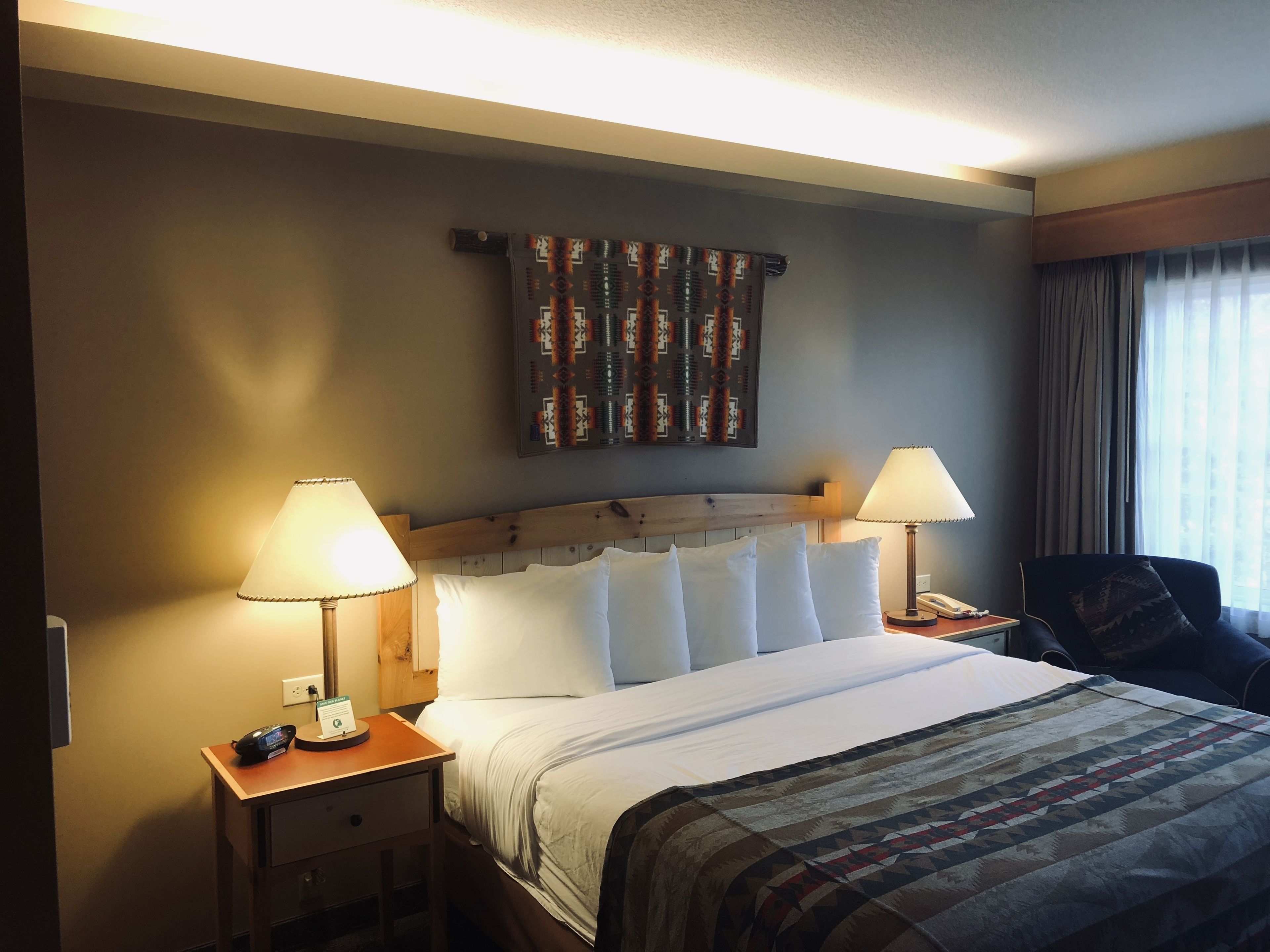 A hotel room with a wooden headboard, a large bed with white linens, two bedside tables with lamps, a telephone, and a chair.