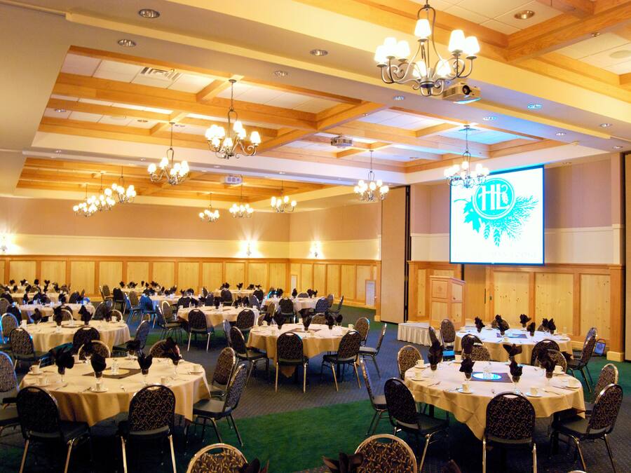 A conference room with multiple round tables and chairs, arranged in a U-shape, with a large screen displaying a logo.