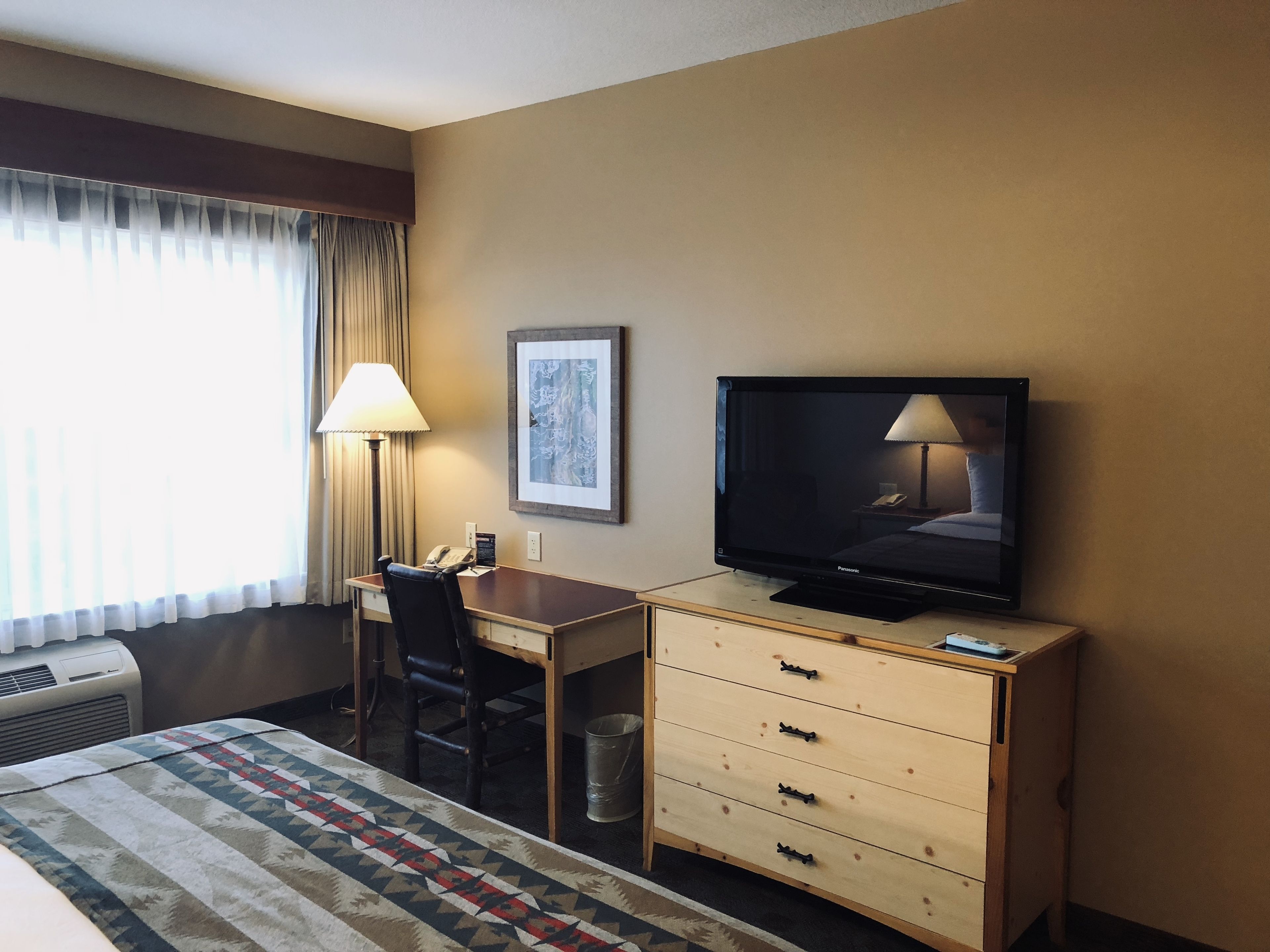 A hotel room with a bed, a desk with a chair, a television, a lamp, and a framed picture on the wall.