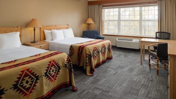 Habitación de hotel con dos camas, un escritorio y una silla. Cuenta con una ventana con cortinas, un calefactor y una lámpara en cada cama.