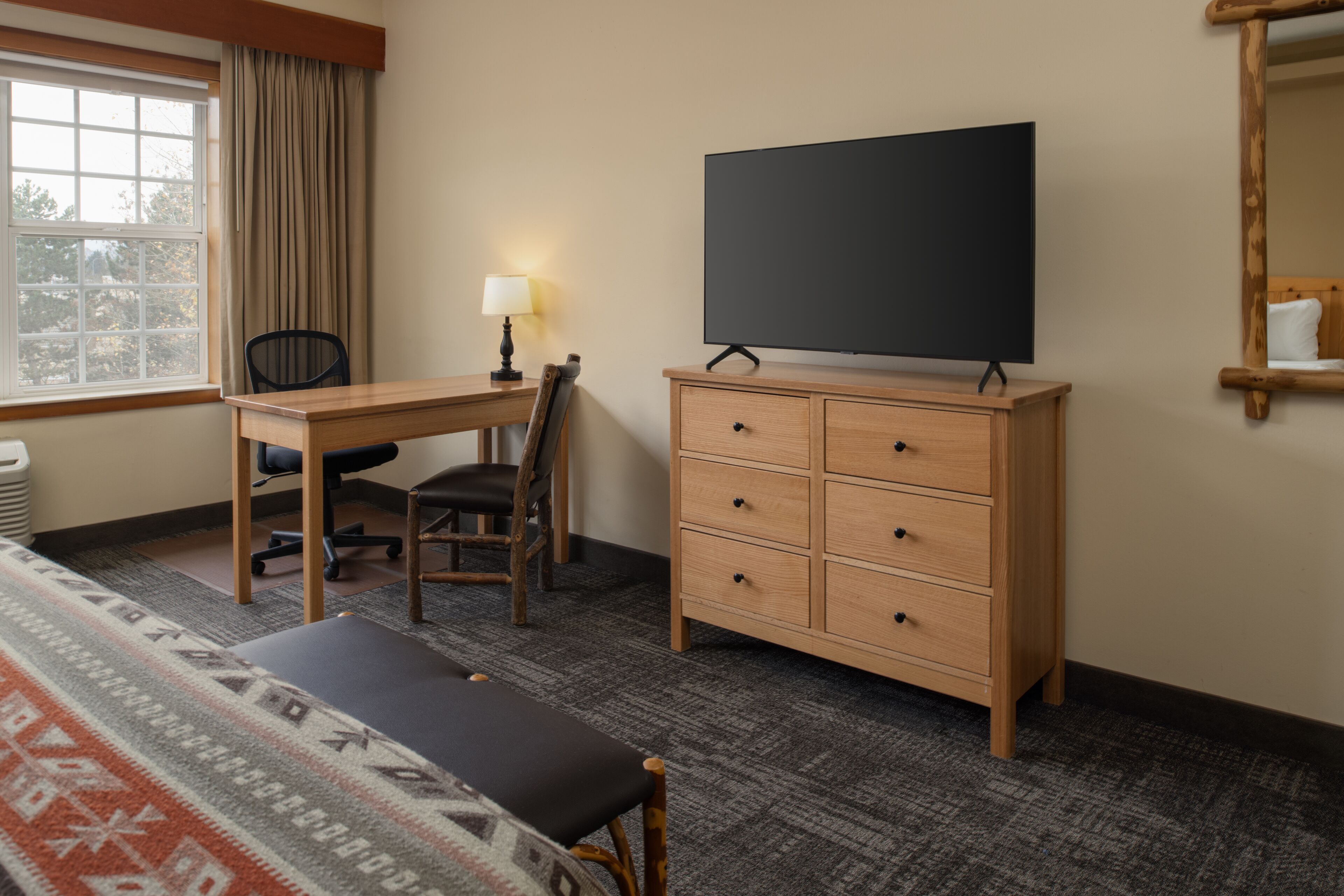 Ein Hotelzimmer mit Holzschreibtisch, einem Sessel, einem Fernseher auf einem Schrank, einem Bett mit gemustertem Überwurf und einem Fenster mit Vorhängen.