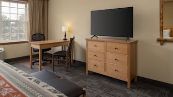 Ein Hotelzimmer mit Holzschreibtisch, einem Sessel, einem Fernseher auf einem Schrank, einem Bett mit gemustertem Überwurf und einem Fenster mit Vorhängen.