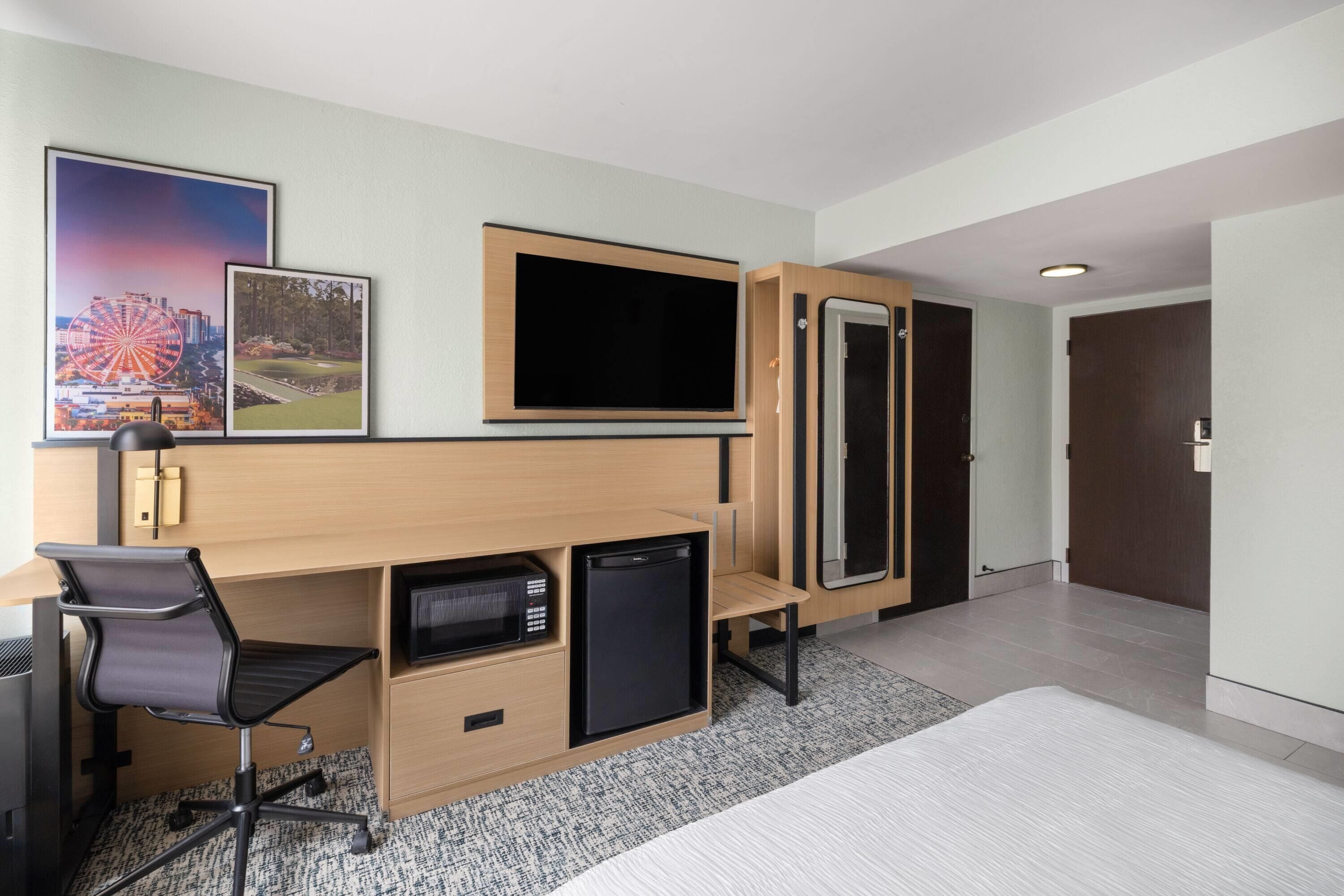 Habitación de hotel con escritorio, silla, televisor, refrigerador pequeño y una cama.