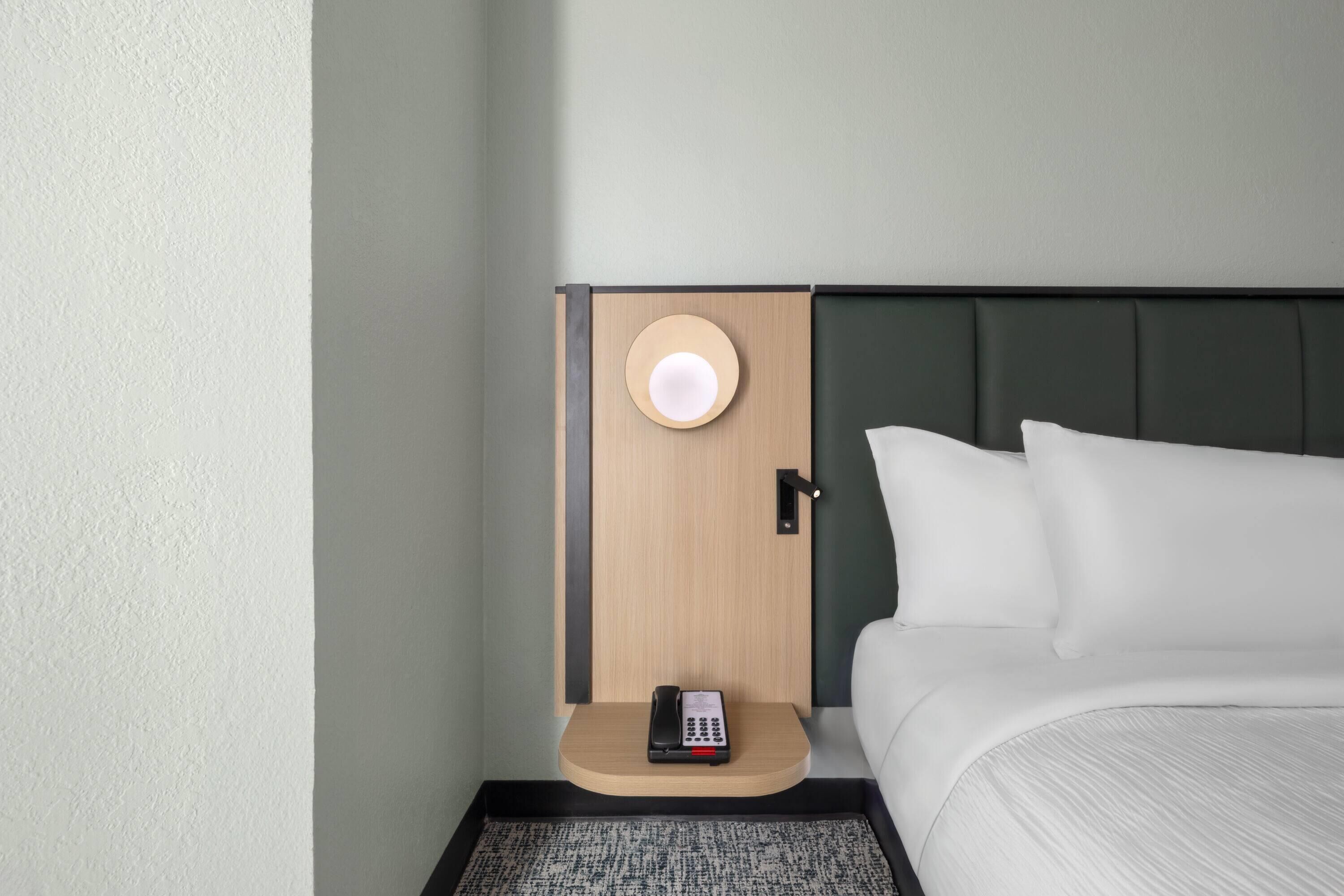 A hotel room with a bed, a nightstand with a telephone, and a door with a round light.