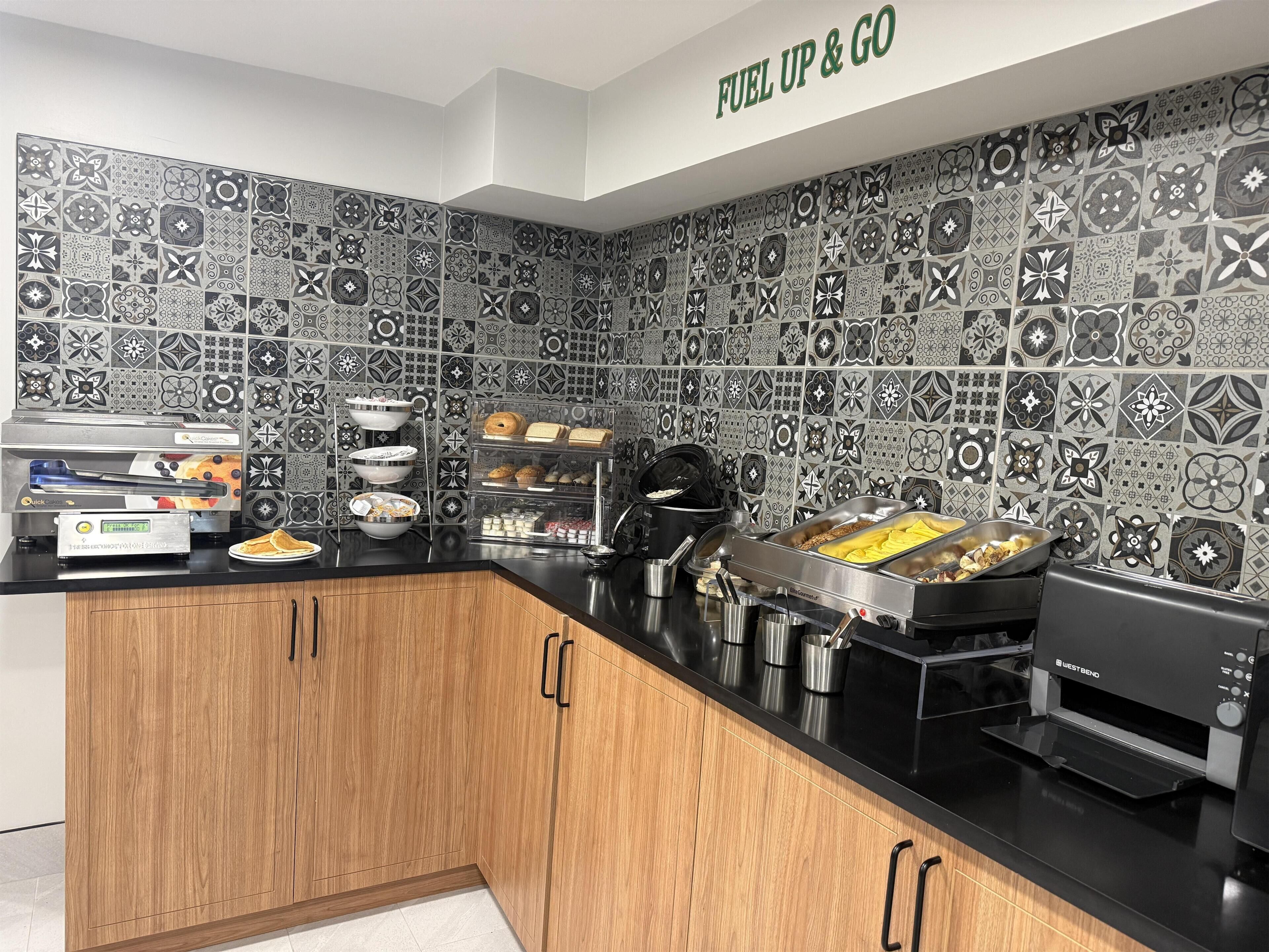 Une cuisine avec un plan de travail noir, des armoires en bois, un grille-pain West Bend et un mur recouvert de carreaux à motifs.