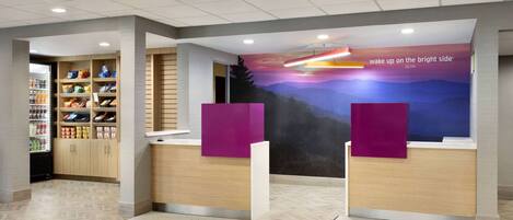 A hotel reception area with a modern design, featuring a reception desk, a wall with a scenic mural, and a shelving unit with snacks and beverages.