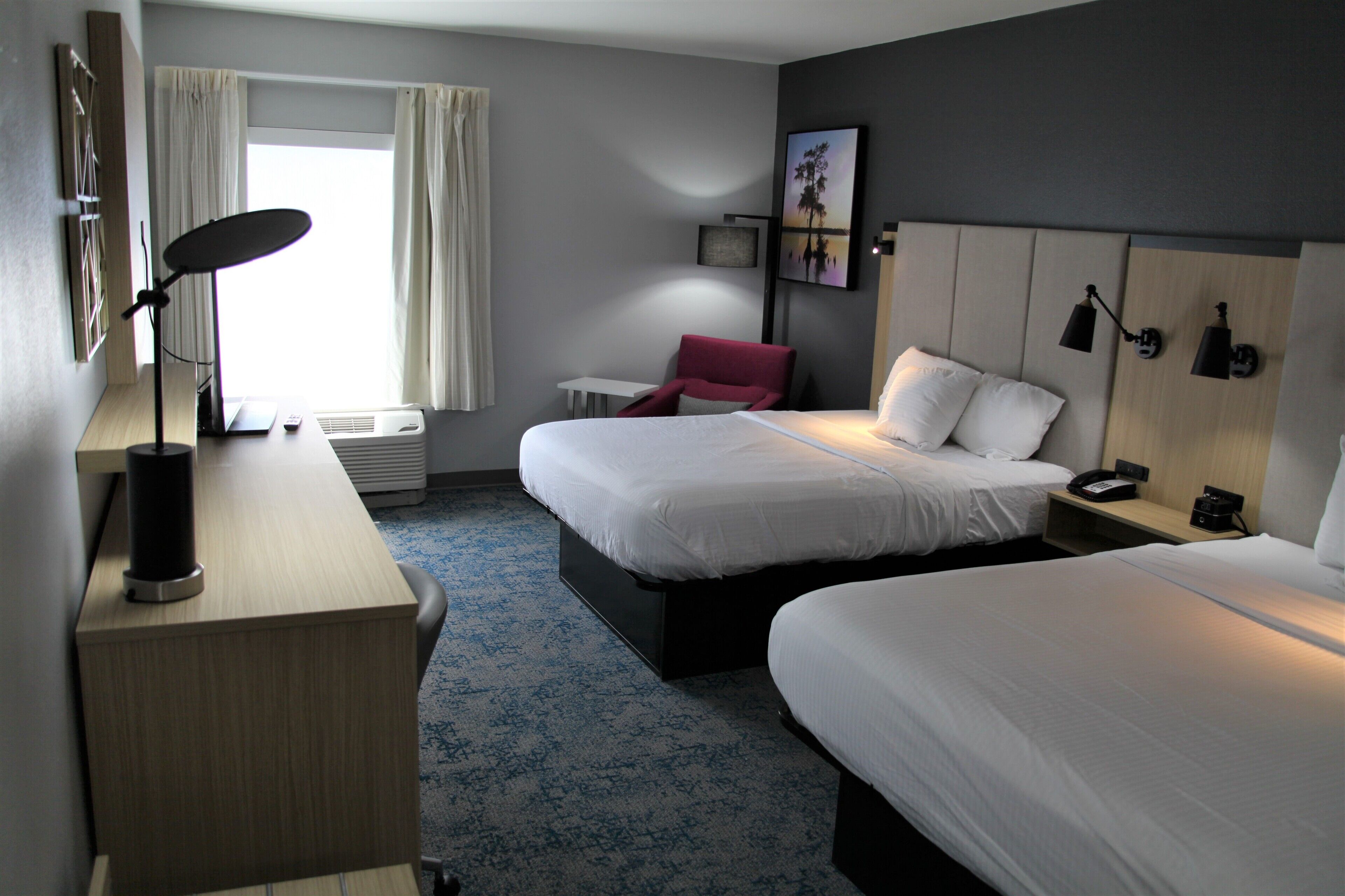 A hotel room with two beds, a desk with a lamp, a chair, a window with curtains, and a painting on the wall.