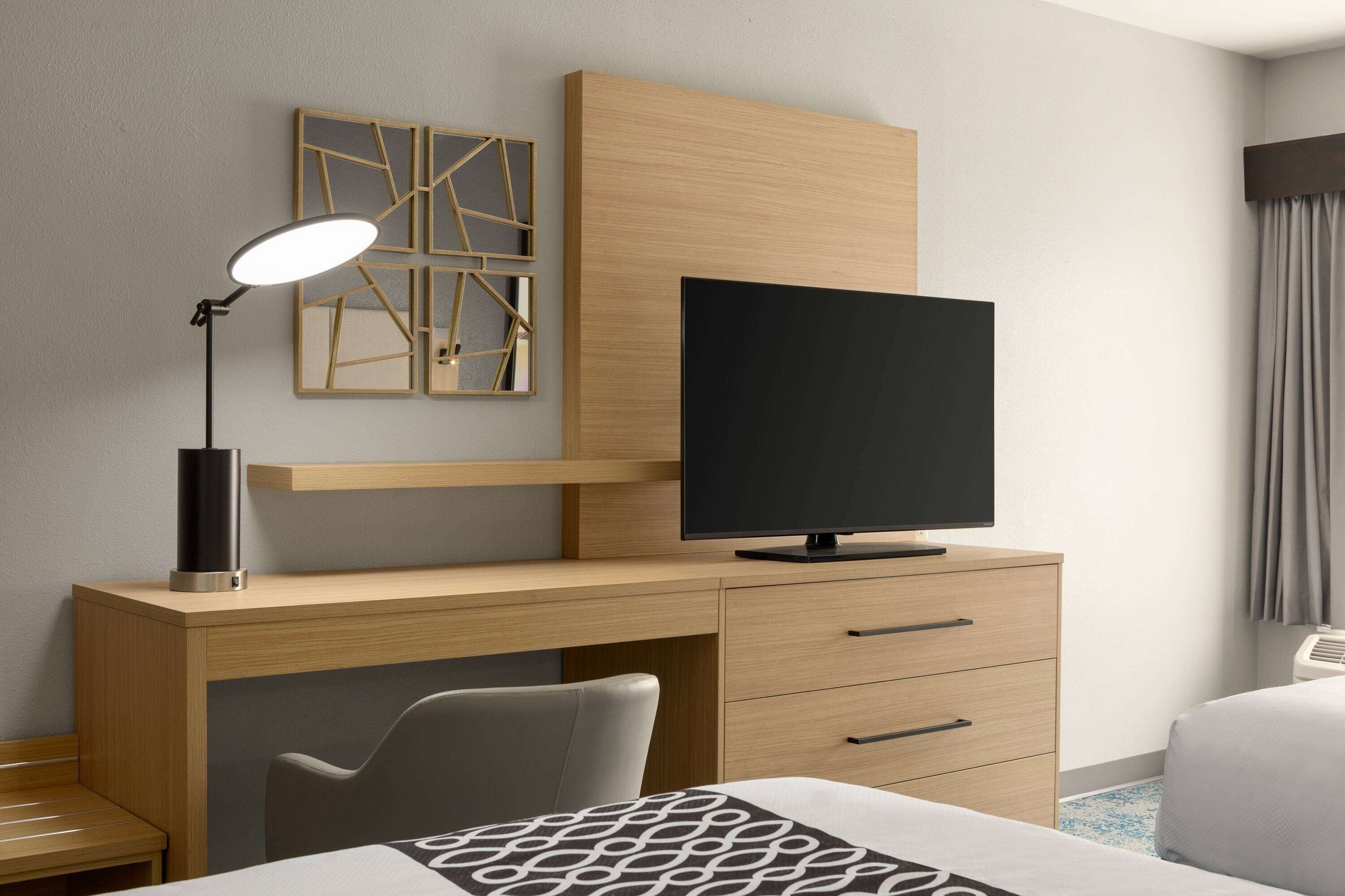 Una habitación de hotel moderna con un escritorio de madera, un televisor de pantalla plana, una lámpara de escritorio, una silla y una cama con colcha estampada.